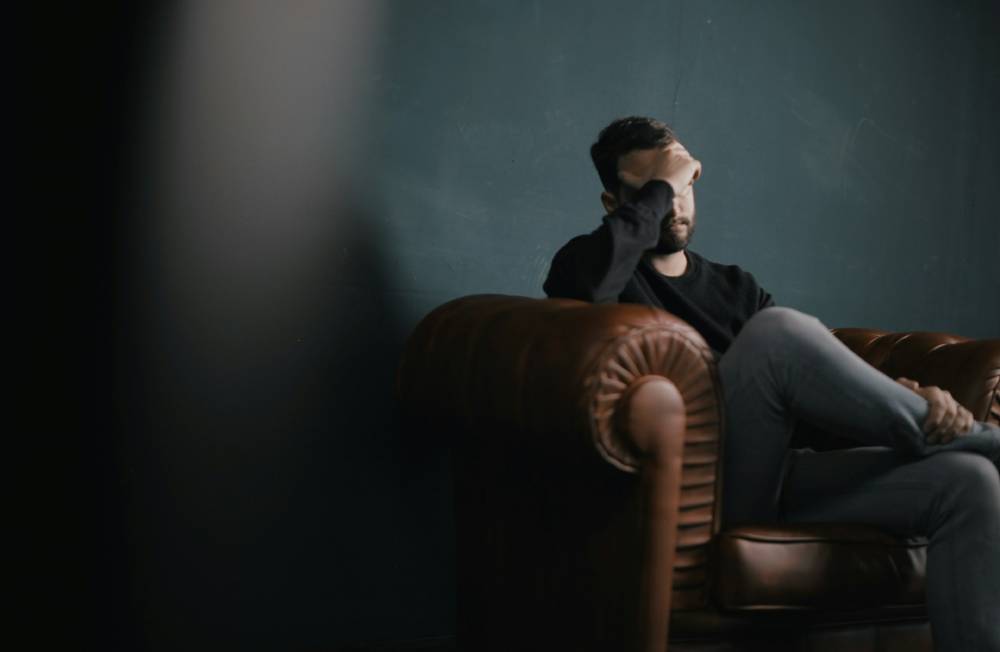uomo seduto su divano con una mano sulla fronte che prova stress