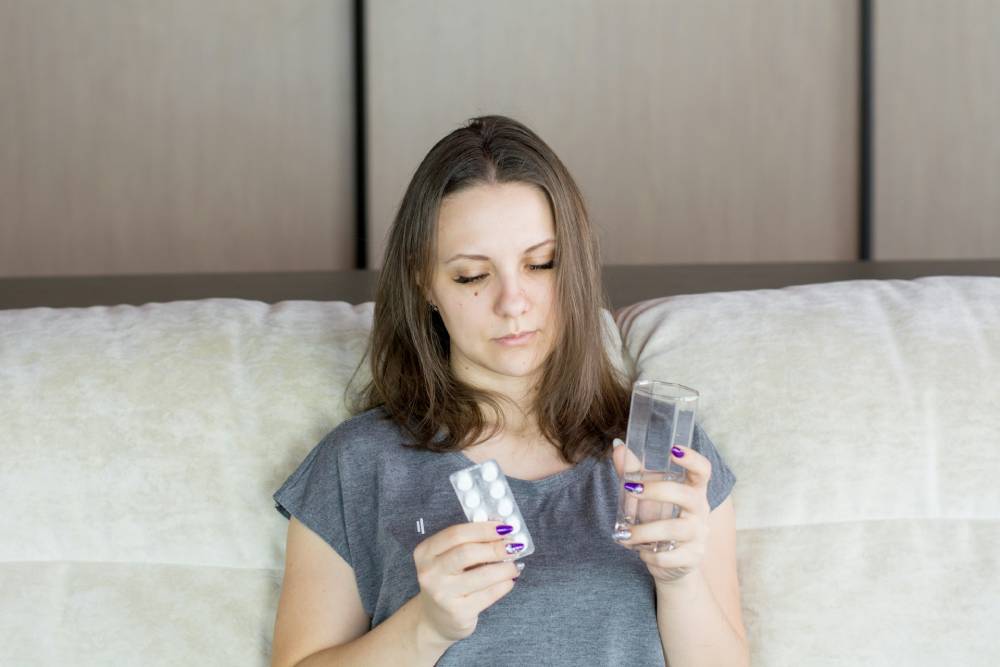 donna seduta su divano bianco con in mano un bicchiere di acqua e una confezione di pastiglie